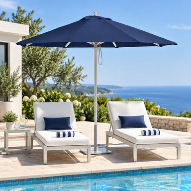 A navy blue patio umbrella stands between two white outdoor chaise lounge chairs with navy and white striped pillows on a sunlit poolside terrace. The background features a clear blue ocean, sea walls, and greenery.